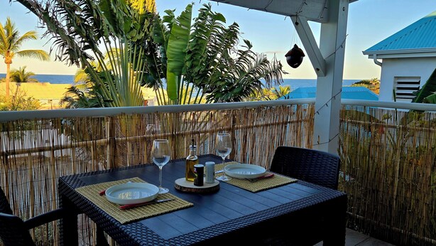 A table set for two with a view of the ocean, a bamboo fence, and a building with a blue roof. - Incredible sea view apartment 🌊 - Swimming pool - Terrace - (Saint-Martin)