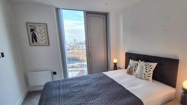 A modern bedroom with a large bed, a nightstand, a window with a city view, and a radiator. - Elegant Skyline Apartment with Exclusive Amenities in the Heart of M3 (Manchester)