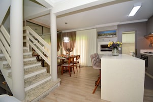 Una cocina moderna con una isla central, un comedor con sillas de madera y una escalera con barandilla de cristal.