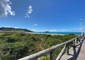 A wooden boardwalk leading to a beach with a clear view of the ocean and distant hills. - Casa Elegante a 100 metros do Mar (Governador Celso Ramos)