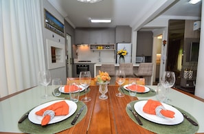 A dining area with a wooden table set for four, a vase of flowers, and a kitchen in the background. - Casa Elegante a 100 metros do Mar (Governador Celso Ramos)