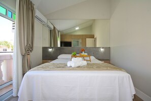 A hotel room with a bed, a bedside table with a vase and fruit, a mirror, and a view of the outside.