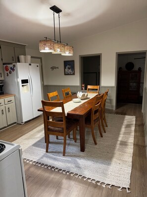 Un comedor con una mesa de madera, sillas, un tapete blanco, una lámpara colgante y un refrigerador.