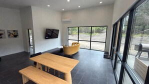 A modern interior with a wooden table and benches, a yellow armchair, a mounted TV, and large windows offering a view of the outdoors.