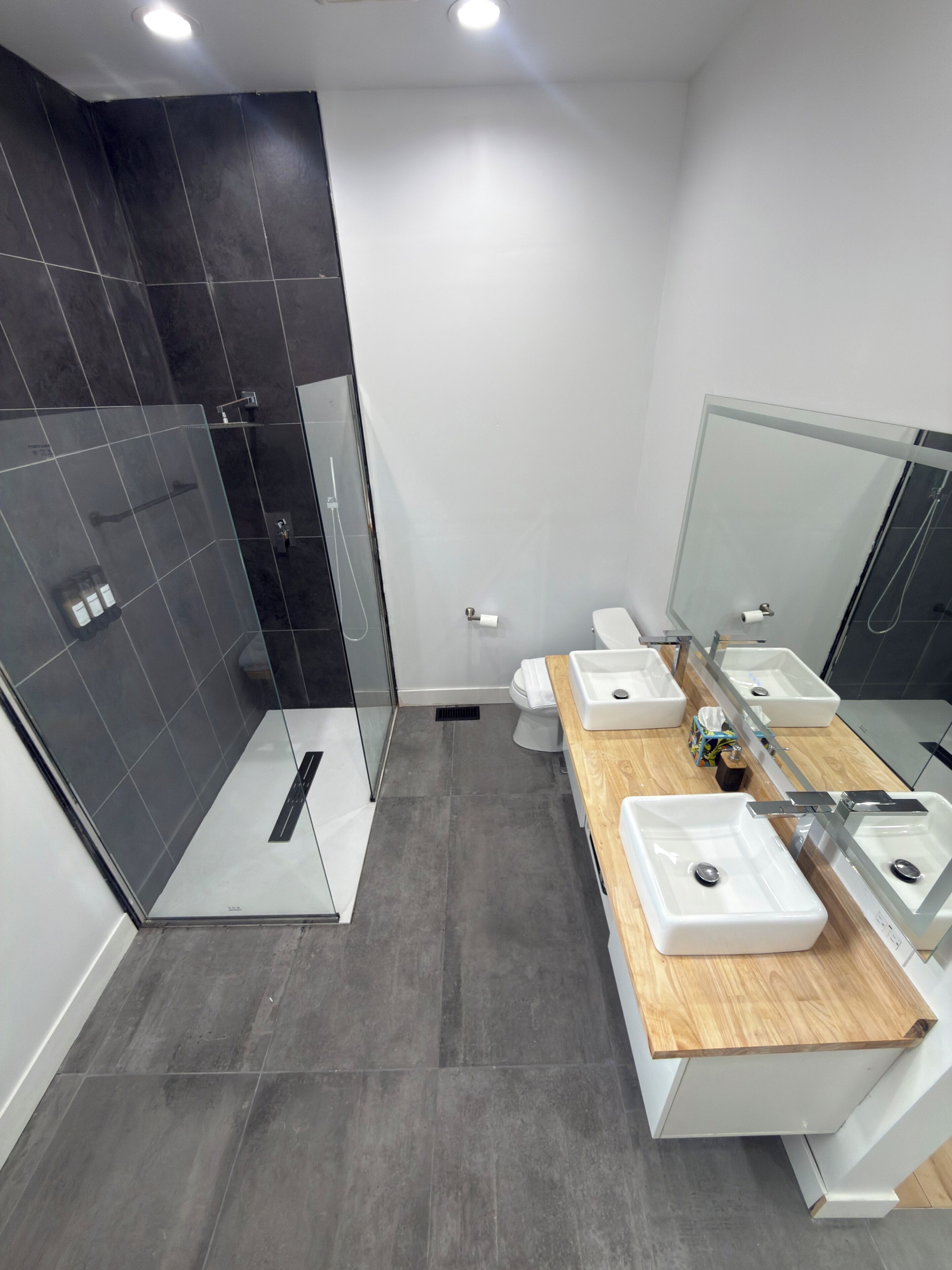 Een moderne badkamer met een douchecabine van glas, twee wastafels met houten aanrechtbladen en een grote spiegel.