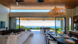 Un intérieur moderne comprenant un coin repas, un canapé et de grandes fenêtres offrant une vue sur l’océan.