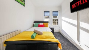 Ein Schlafzimmer mit Bett, Kissen, Decke, einem Gemälde, einem Fenster und einem Schild mit der Aufschrift 'NETFLIX'.