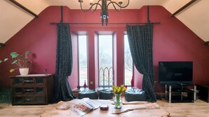 A loft-style room with a sloped ceiling, a chandelier, a television, a wooden table, and a large window with curtains. - Kaber Chapel (Kirkby Stephen)