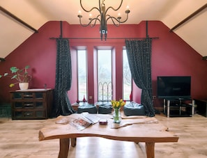 A loft-style room with a sloped ceiling, a chandelier, a television, a wooden table, and a large window with curtains. - Kaber Chapel (Kirkby Stephen)