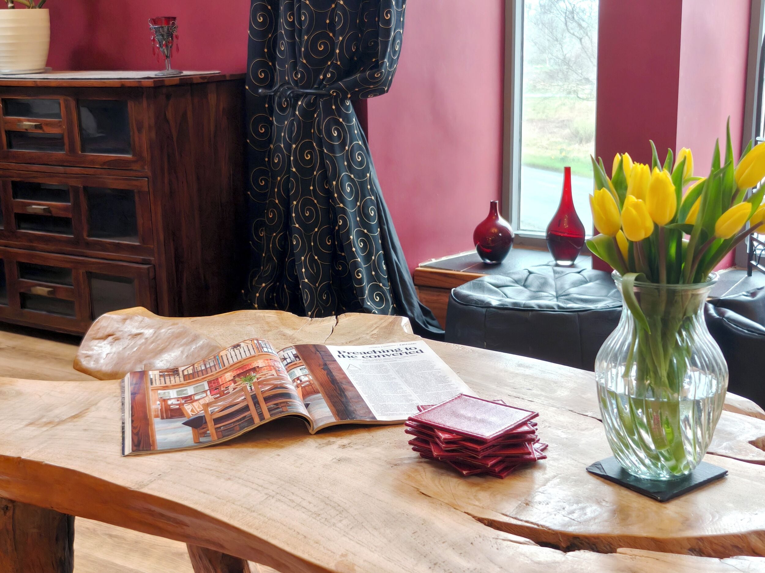 Een houten tafel met een tijdschrift, een stapel rode kaarten en een vaas met gele bloemen.