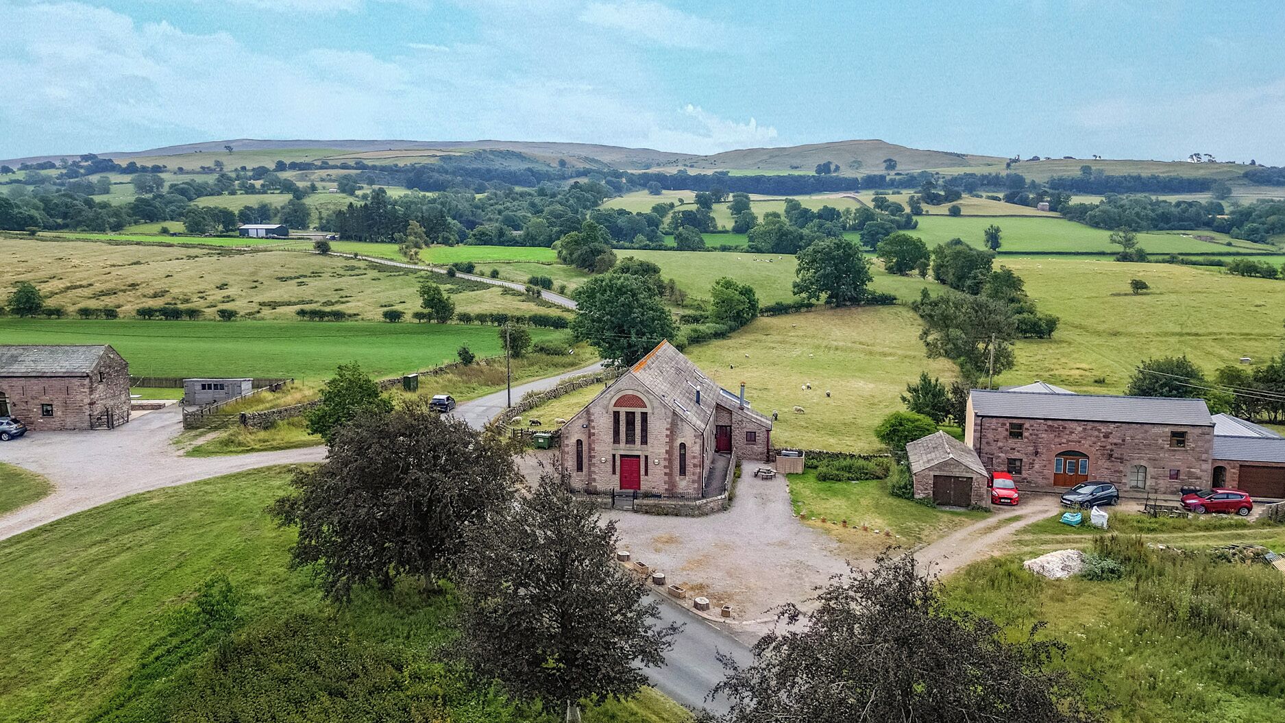 Paisaje rural con un edificio de techo rojo destacada, posiblemente una iglesia, rodeado de campos verdes y algunas casas dispersas.