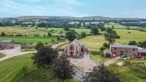 Paisaje rural con un edificio de techo rojo destacada, posiblemente una iglesia, rodeado de campos verdes y algunas casas dispersas.