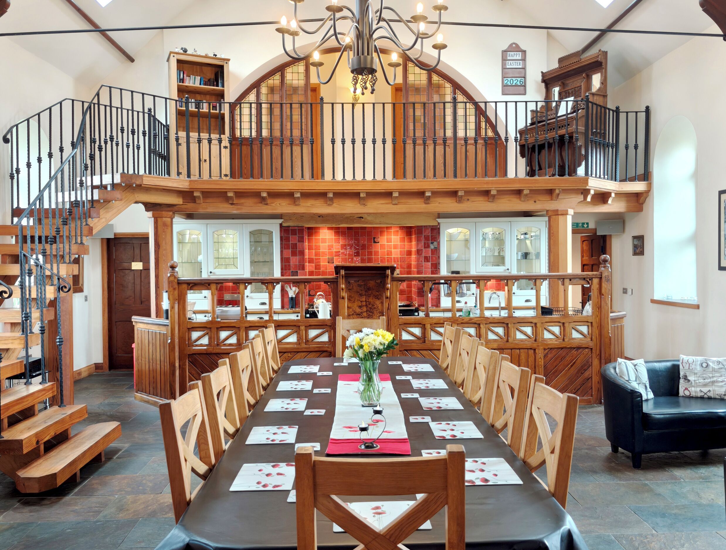 Un comedor con una larga mesa dispuesta para una comida, sillas de madera, un candelabro y una escalera que sube a un nivel superior.