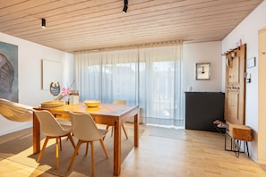 A modern living room with a wooden dining table, two chairs, a hammock, and a large window with sheer curtains. - Eulenschlupf – Black Forest Premium Apartment with Sauna, Home Cinema, Hammock, Terrace (Ühlingen-Birkendorf)