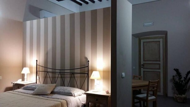 A bedroom with a bed, bedside lamps, a nightstand, a chair, a dining table, and a door. - Antico Portale (Acquasparta)