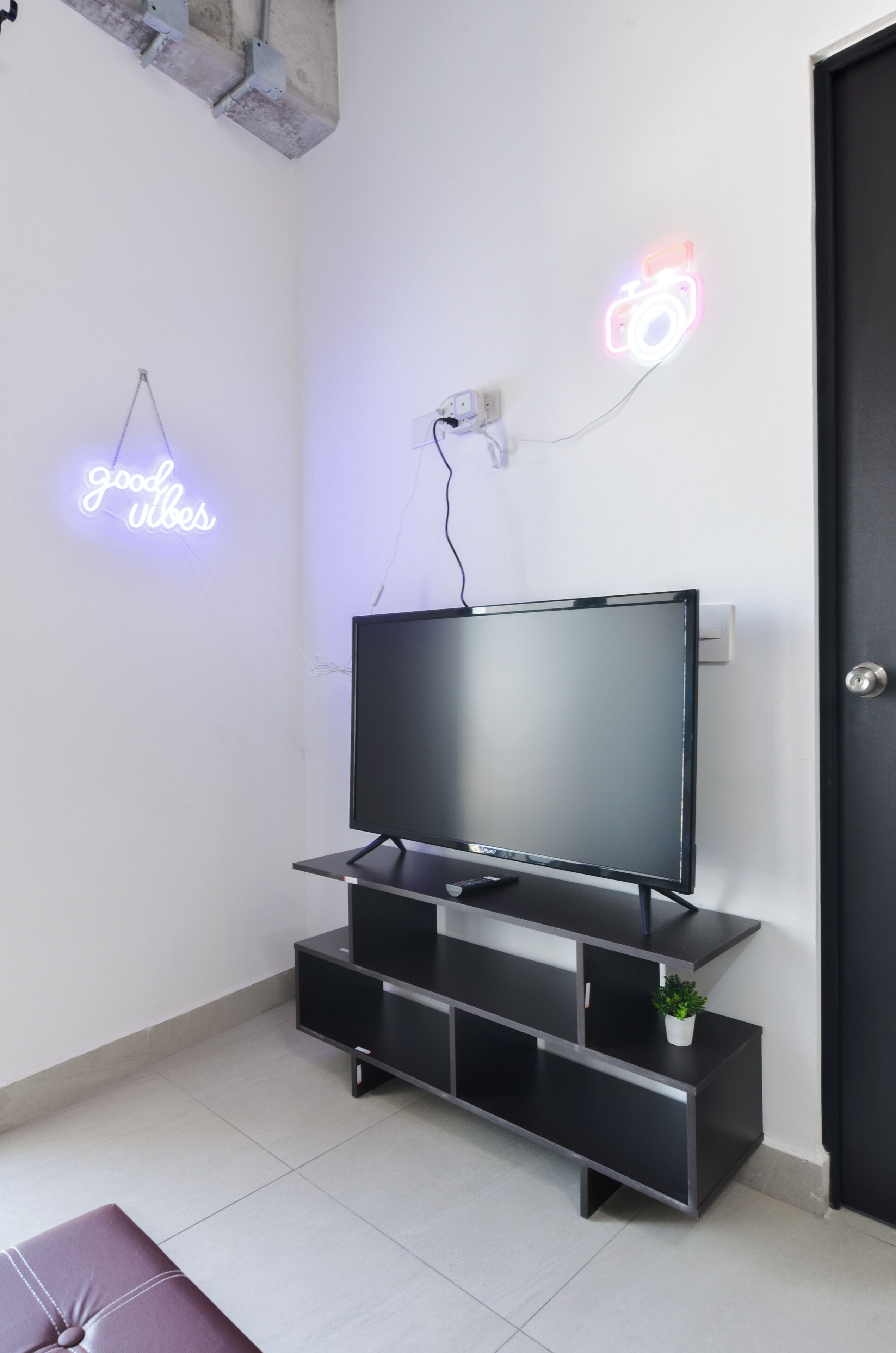 A modern living room with a flat-screen TV on a black stand, a neon sign on the wall, and a door to the right.