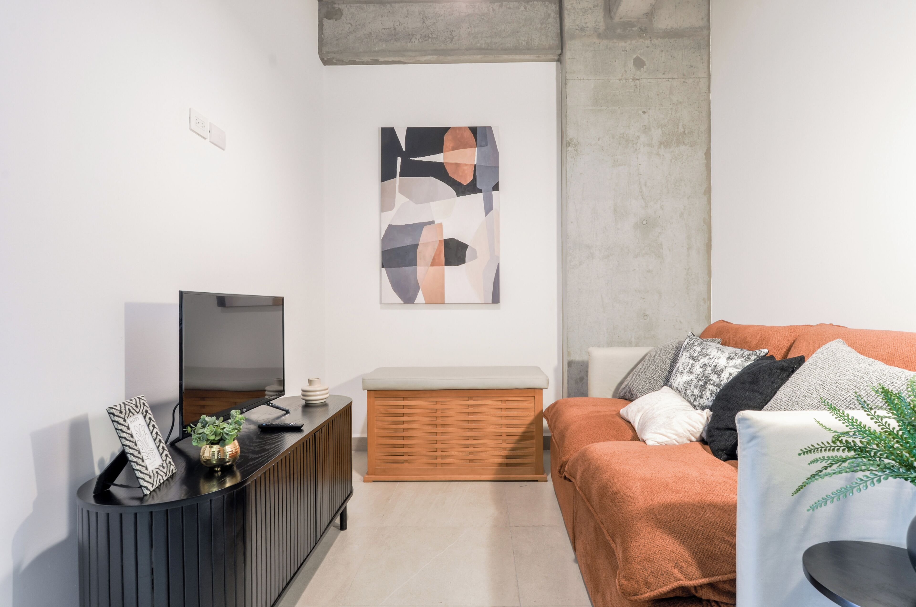 A modern living room with a flat-screen TV, a wooden bench, a sofa with cushions, and a wall-mounted abstract art piece.