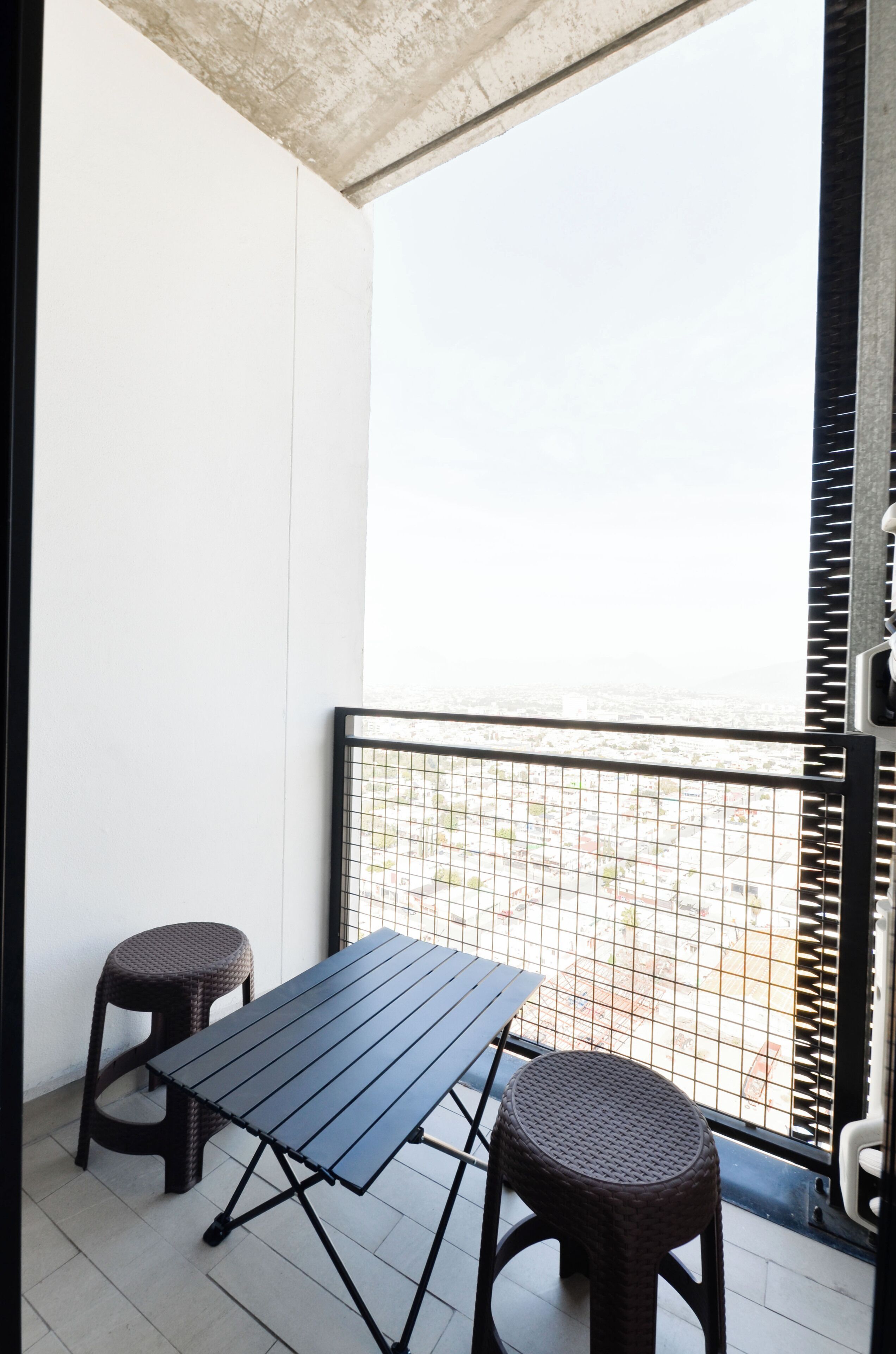 Ein Balkon mit Tisch, zwei Hocker, Metallgeländer und Blick auf die Stadtlandschaft.