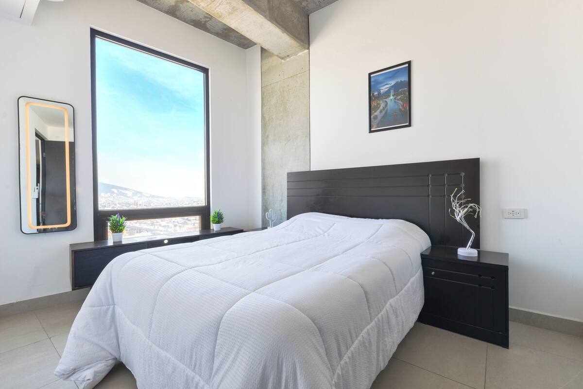 Un dormitorio moderno con un ventanal grande, una cama, una mesita de noche, un espejo y un cuadro en la pared.