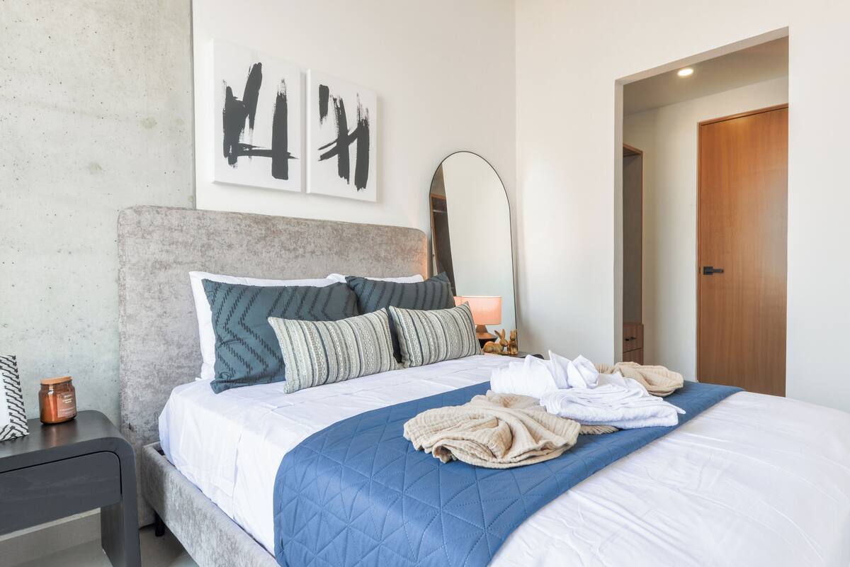 Una habitación de hotel con cama, almohadas, una mesita de noche, lámpara y cuadros en la pared.
