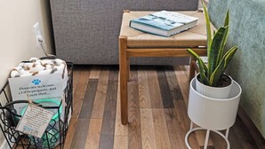 Ein Zimmer mit einem grauen Sofa, einem Holzstuhl mit einem Buch, einem weißen Pflanzenständer und einem Korb mit Hundezubehör.