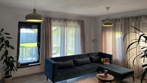 Een woonkamer met een hoekbank, een ronde salontafel en een groot raam met gordijnen.
