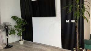 Een moderne woonkamer met een flatscreen televisie op een zwart paneel, een staande ventilator, een houten salontafel en twee potplanten.
