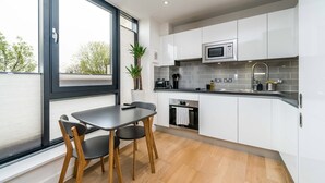 A modern kitchen with white cabinets, a black countertop, and a window with a view of trees. - Stylish Flat by Victoria Park (London)