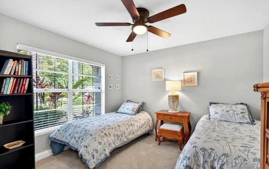 A bedroom with two beds, a ceiling fan, a bookshelf, and a window with blinds.