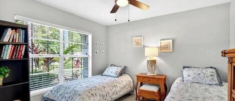A bedroom with two beds, a ceiling fan, a bookshelf, and a window with blinds.