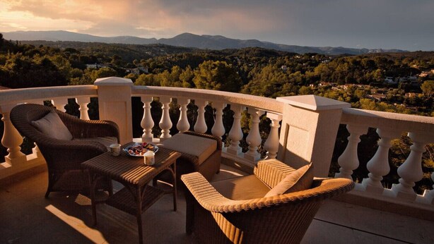 Un balcon agrémenté de meubles en osier, une table avec une assiette de nourriture, et une vue sur une ville et des montagnes au loin.