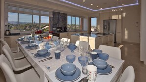 Un espace de restauration moderne avec une longue table dressée pour un repas, de grandes fenêtres et une cuisine en arrière-plan.