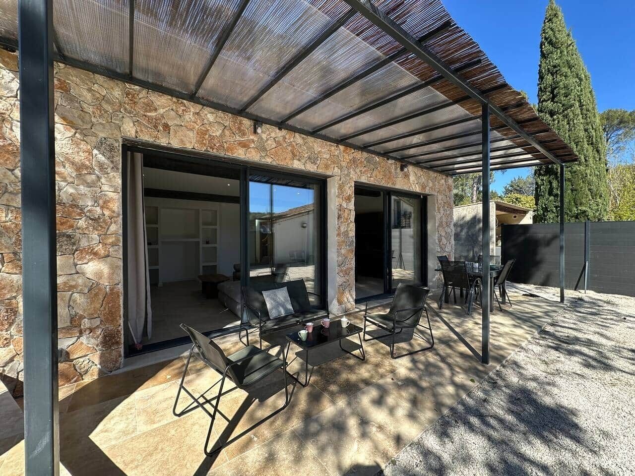 Une terrasse couverte, aménagée avec du mobilier d’extérieur, des murs en pierre et offrant une vue sur l’intérieur.