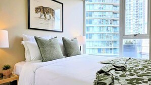 Une chambre d’hôtel avec un lit, une reproduction d’une image d’animal encadrée au mur, une fenêtre donnant sur un immeuble, et une table de chevet avec une lampe et une petite plante.