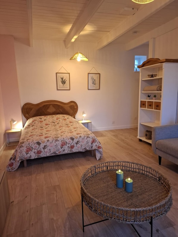 Une chambre avec un lit en bois, une petite table ronde en osier décorée de bougies, une table de chevet, une chaise bleue et une étagère blanche.