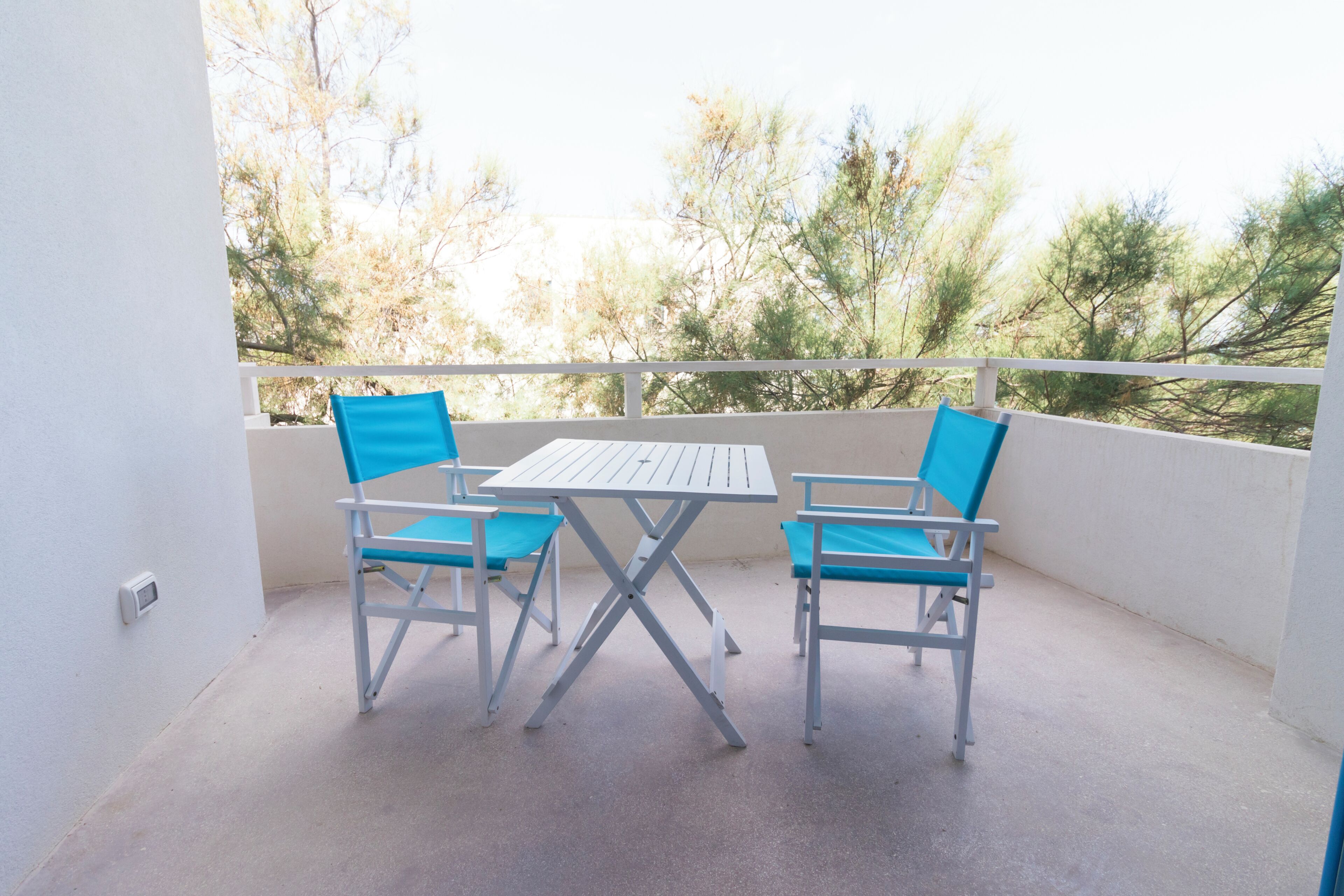 Un balcon avec une table blanche et des chaises bleues, donnant sur des arbres.
