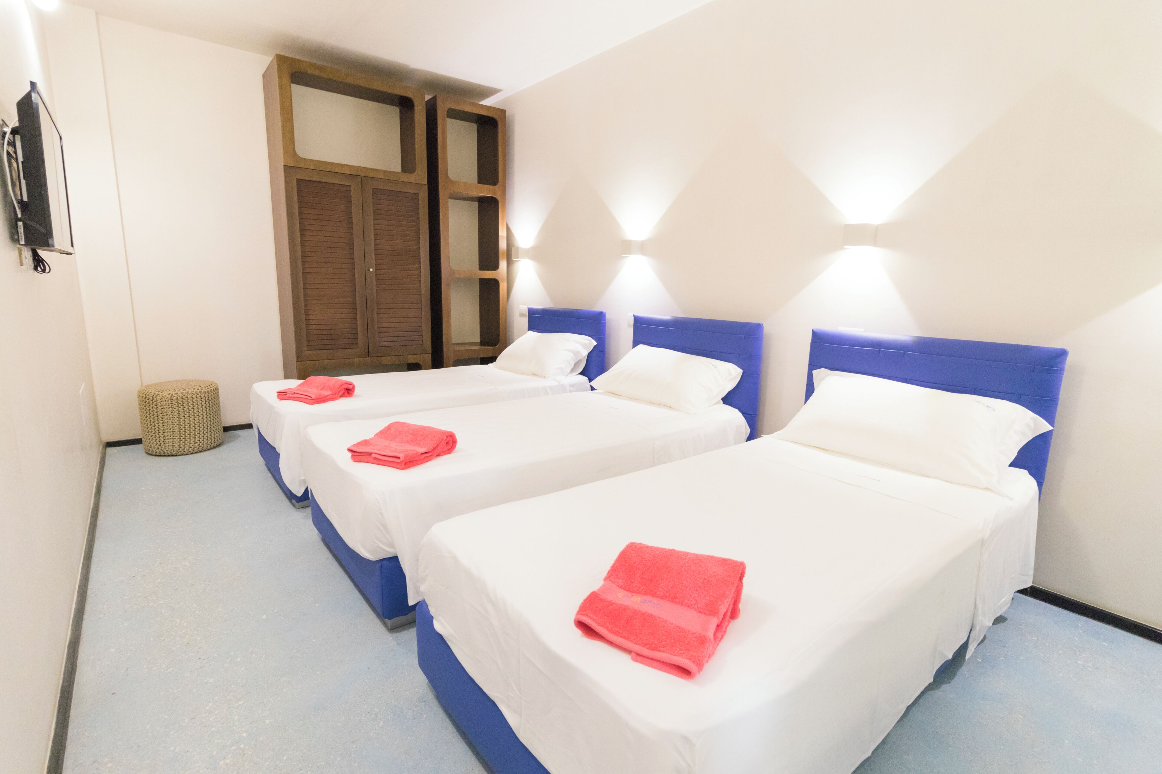 A hotel room with two beds, each with a blue headboard and white bedding, and a small round stool.