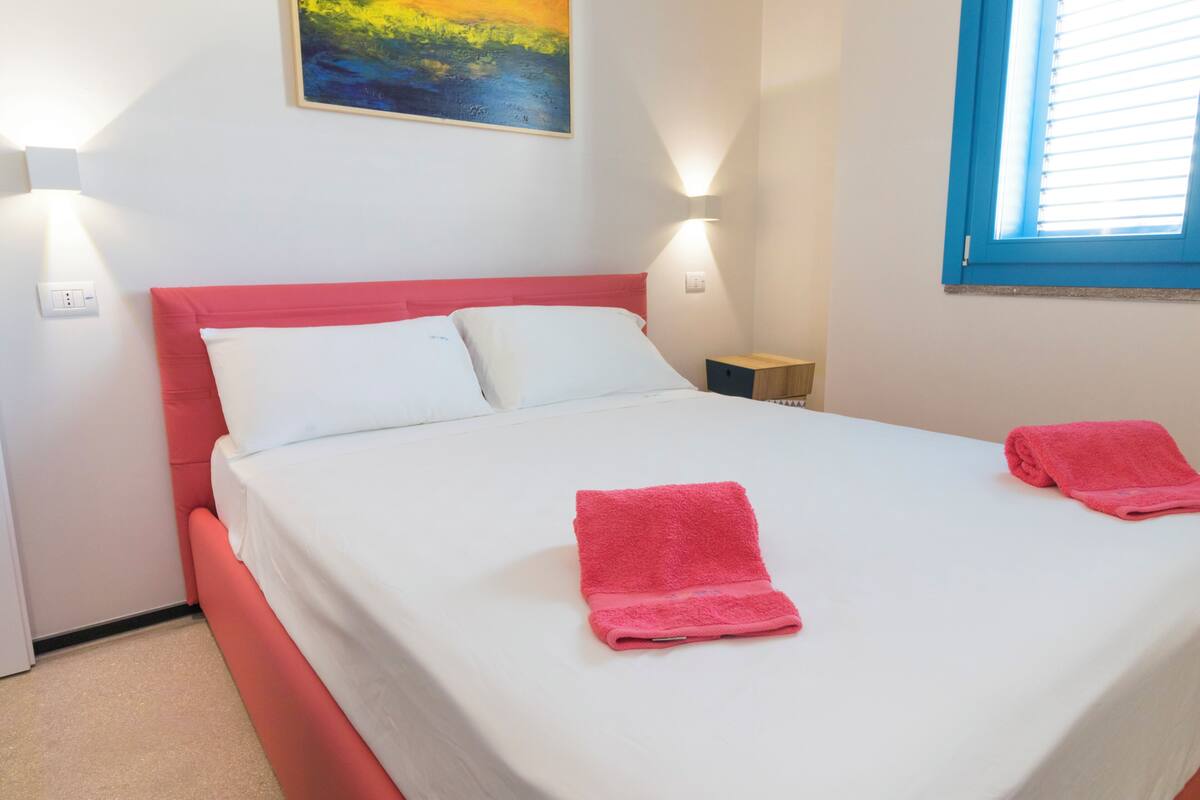 A bed with white bedding and pink pillows, two pink towels, a painting on the wall, and a window with blinds.