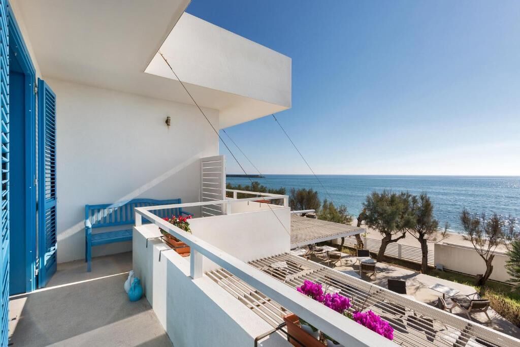 A balcony with a view of the sea, a blue bench, and outdoor furniture.