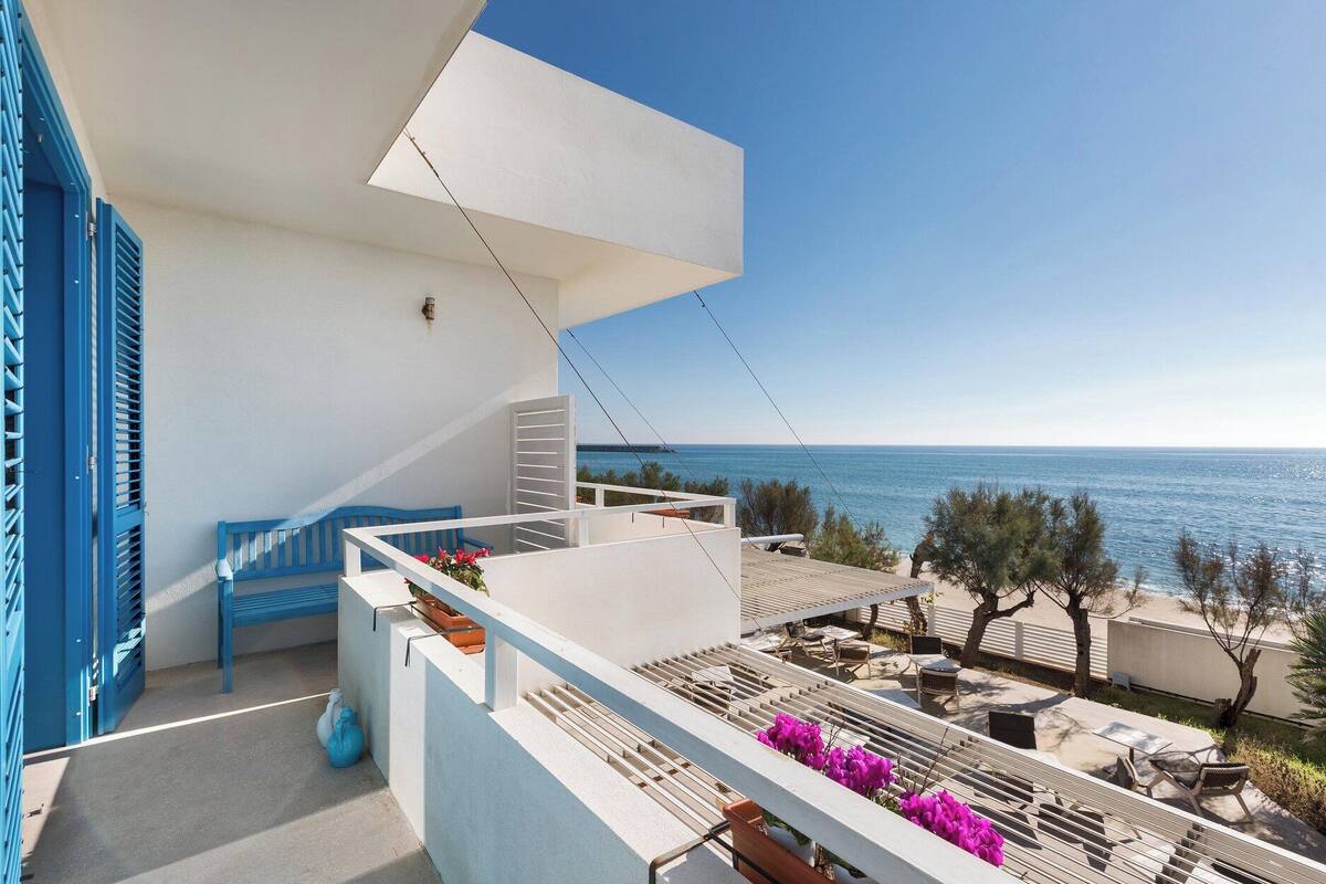 A balcony with a view of the sea, a white building, and blue shutters.