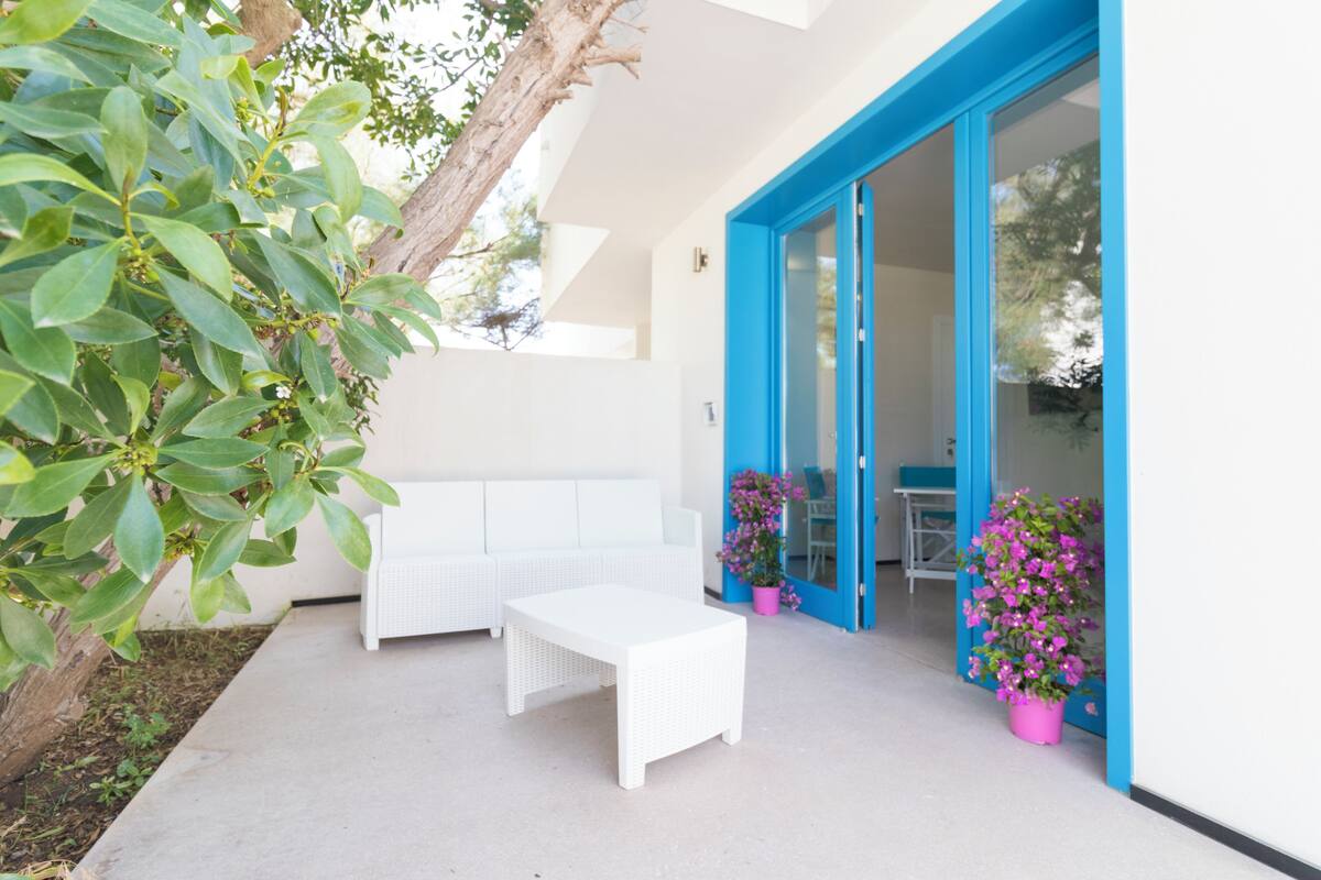A modern outdoor seating area with a white table and bench, surrounded by potted plants and a blue-framed door.