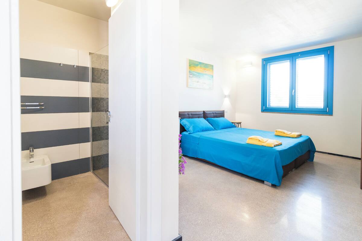 A bedroom with a bed, a window, a bathroom with a sink, and a shower area.