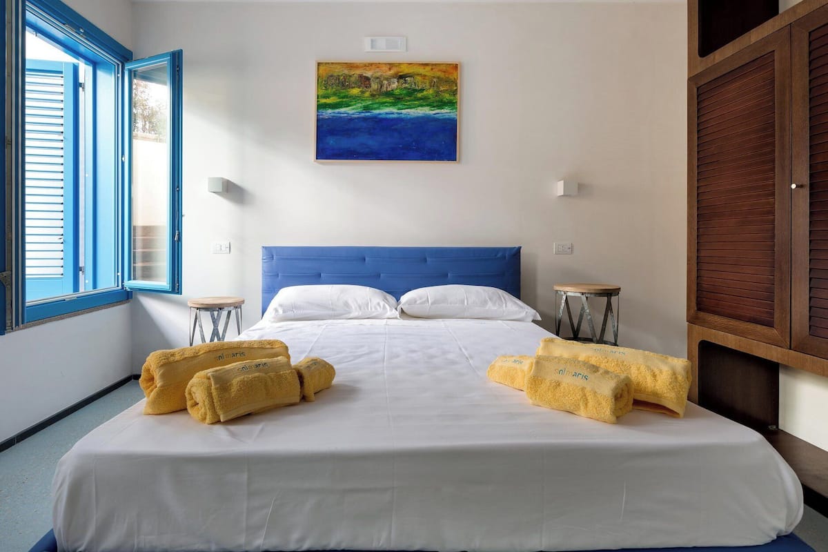 A hotel room with a bed, two bedside tables, a painting on the wall, and a window with blue shutters.