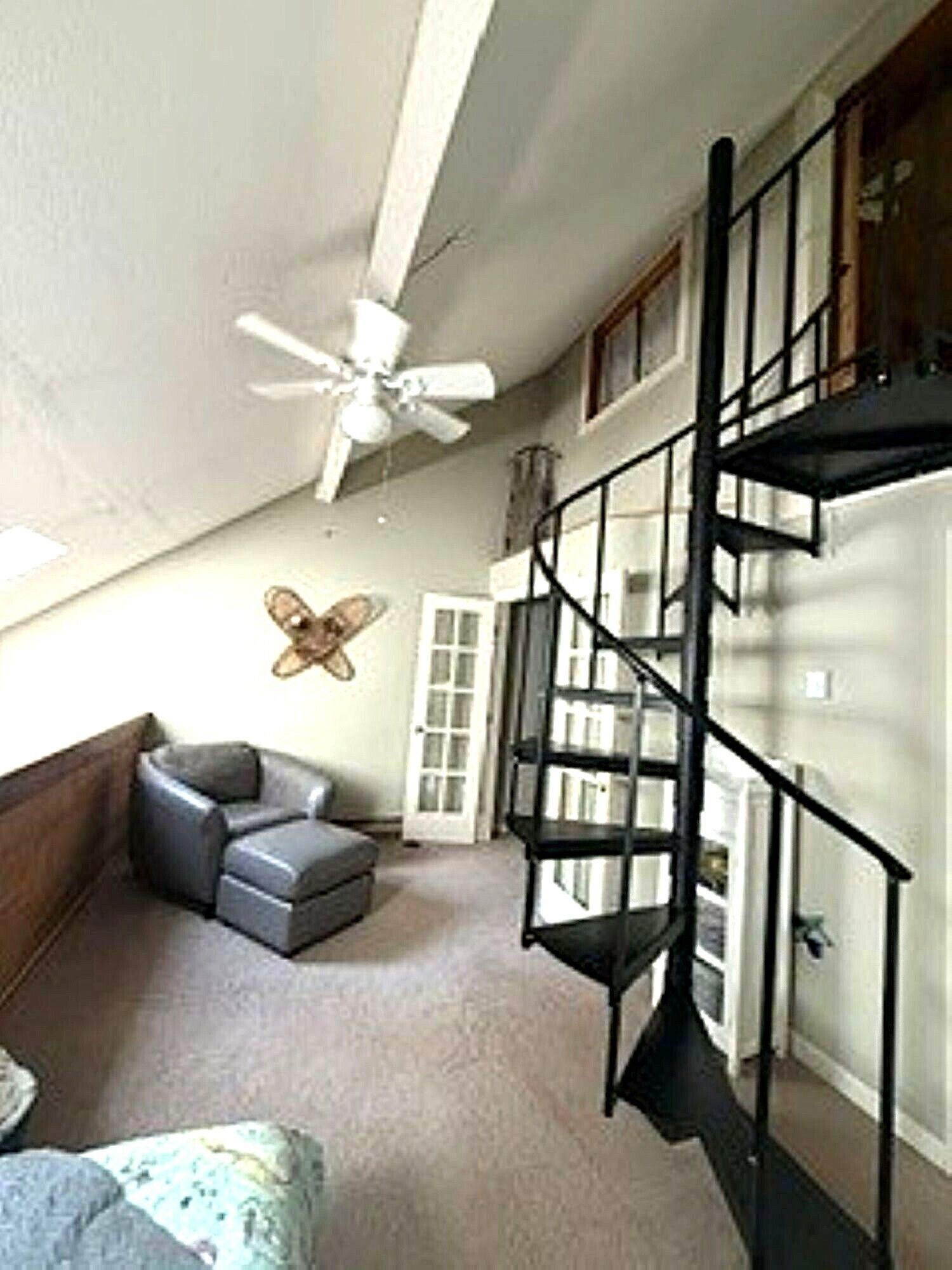 Una habitación con ventilador de techo, sofá gris, reposapiés estampado, escalera en espiral negra y decoración de mariposa en la pared.