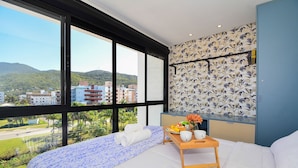 A bedroom with a large window, a bed with a blue patterned bedspread, a wooden bedside table with a tray of fruit and flowers, and a wall with a floral wallpaper. - Charme e Conforto - 400m Praia de Palmas (Governador Celso Ramos)