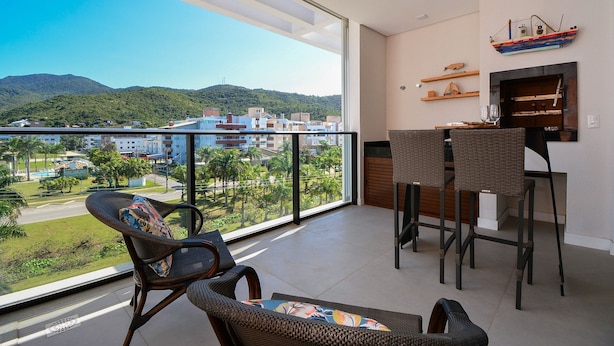 A balcony with wicker furniture, a bar area, and a view of a cityscape and mountains. - Charme e Conforto - 400m Praia de Palmas (Governador Celso Ramos)