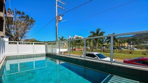 A swimming pool with clear water, surrounded by a glass railing, with a view of palm trees and cars in the background. - Charme e Conforto - 400m Praia de Palmas (Governador Celso Ramos)