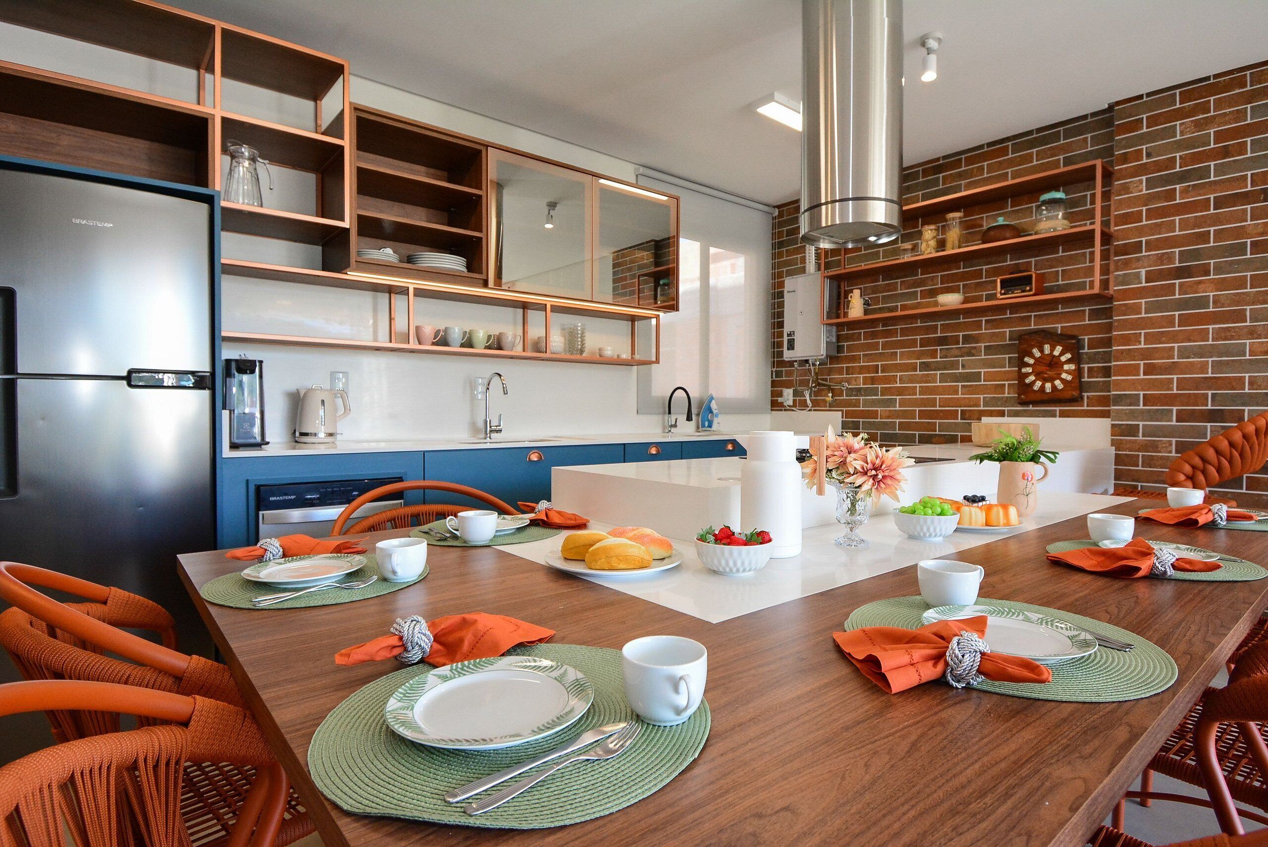 A modern kitchen with a brick wall, wooden shelves, and a dining area set for four with orange chairs and green placemats.