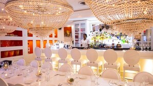 A restaurant with round tables, white chairs, and a bar area with a bartender.