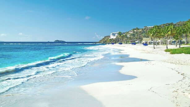 A beach with white sand, ocean waves, and a building with palm trees.